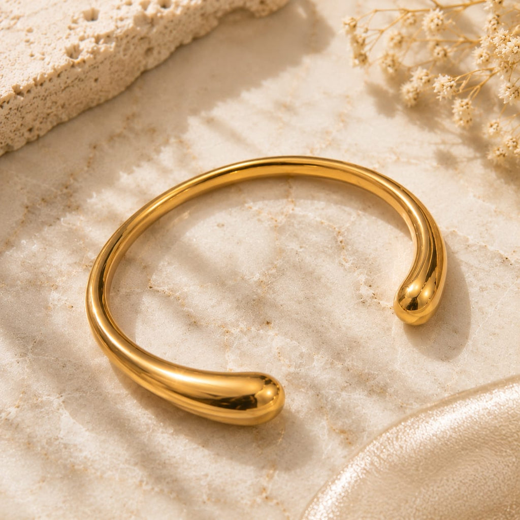 Teardrop ball end open cuff bangle βραχιόλι, χρυσό ανοξείδωτο ατσάλι, flat lay σε travertine — Celestial Steel